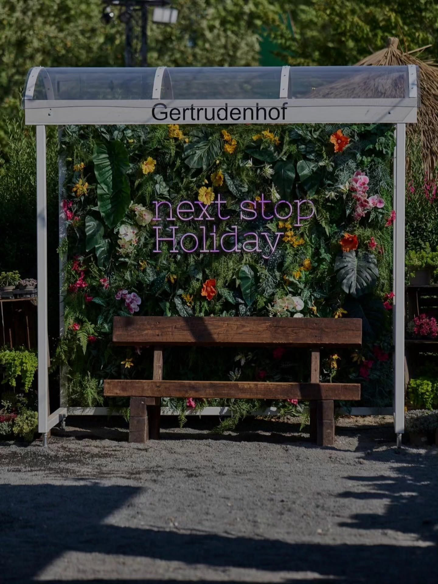 Dekorierte Gertrudenhof-Haltestelle mit üppiger Blumenwand, Neon-Schriftzug next stop Holiday und einer Holzbank